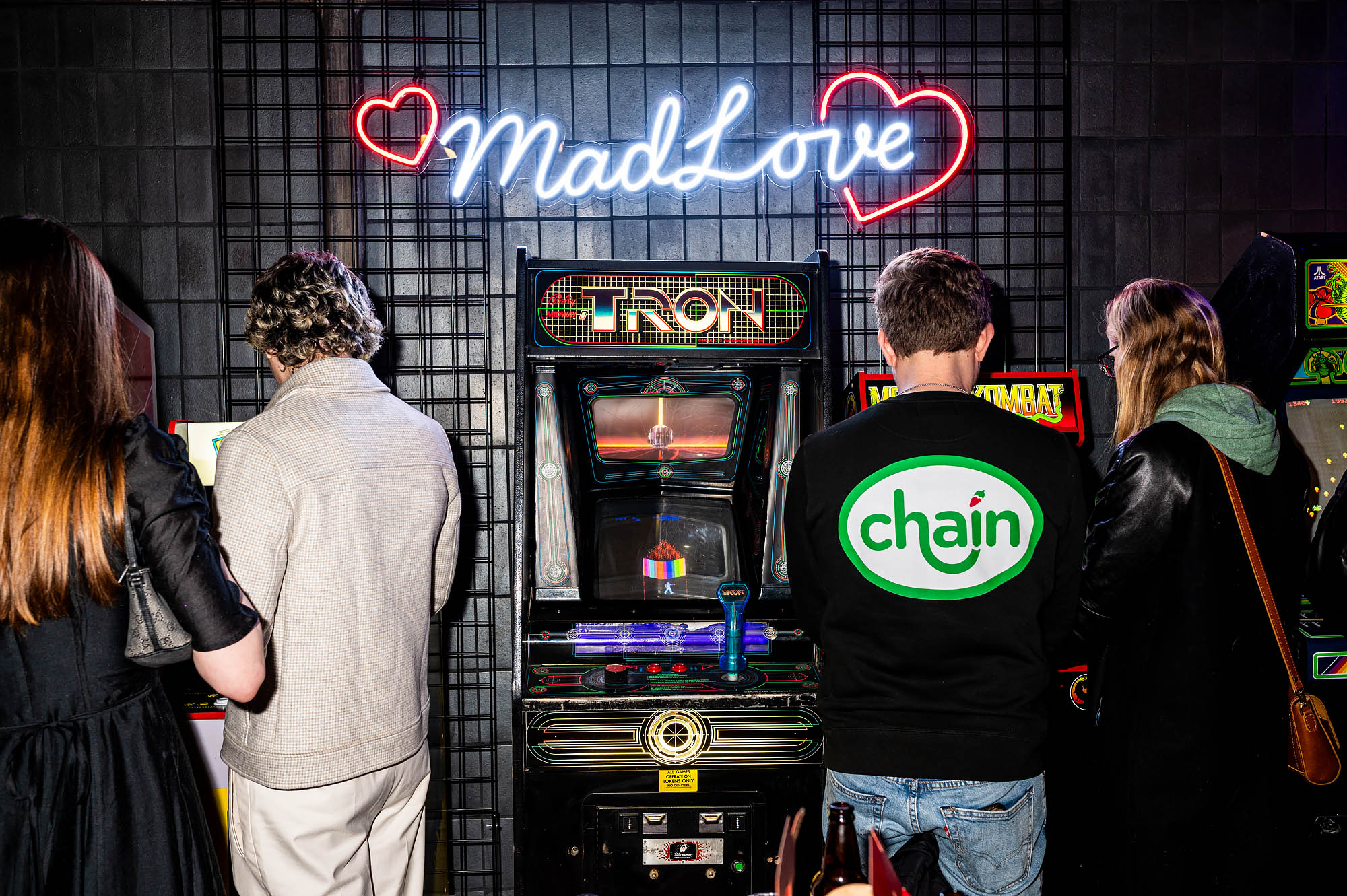 A young man in a Chain jacket stands at an old video game consol next to a Tron video game, with other attendees. 