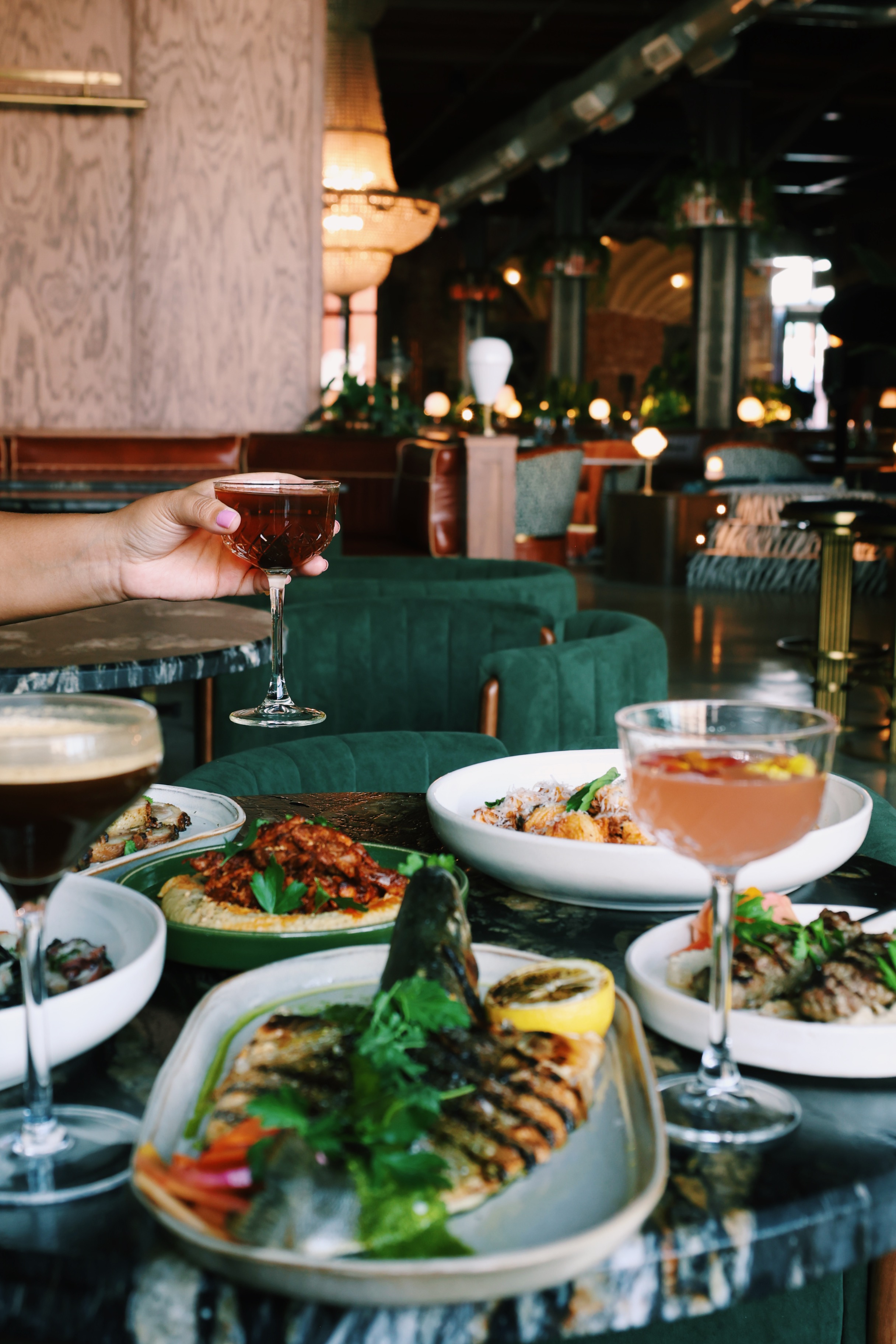 A table is loaded with small plates of Mediterranean-inspired food and cocktails. A hand reaches into the left side of the frame, holding a cocktail. Behind, booths of green velvet and the interior of a restaurant can be seen.