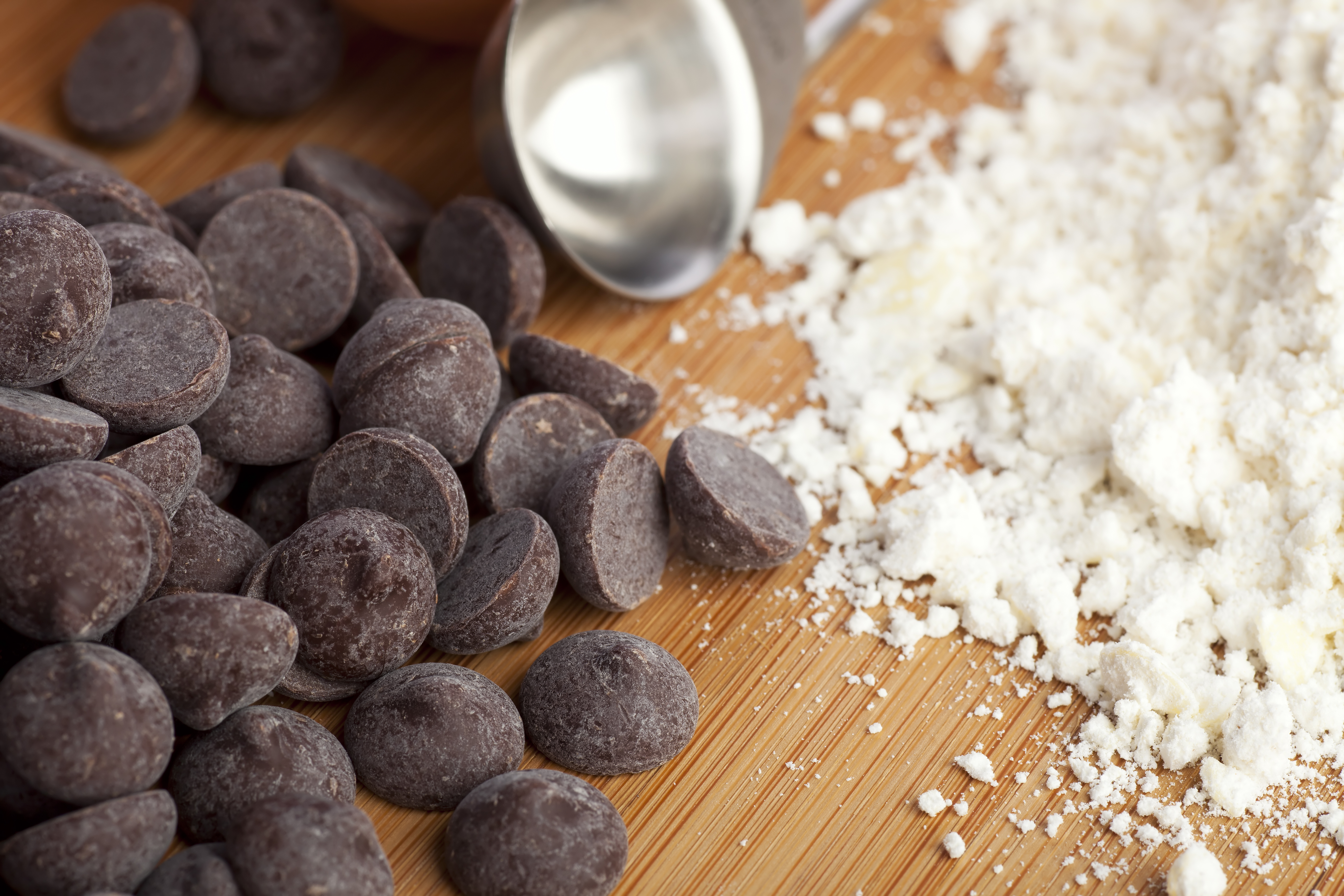 A close-up of a pile of chocolate chips next to a pile of flour, with a metal measuring spoon on a wood surface.