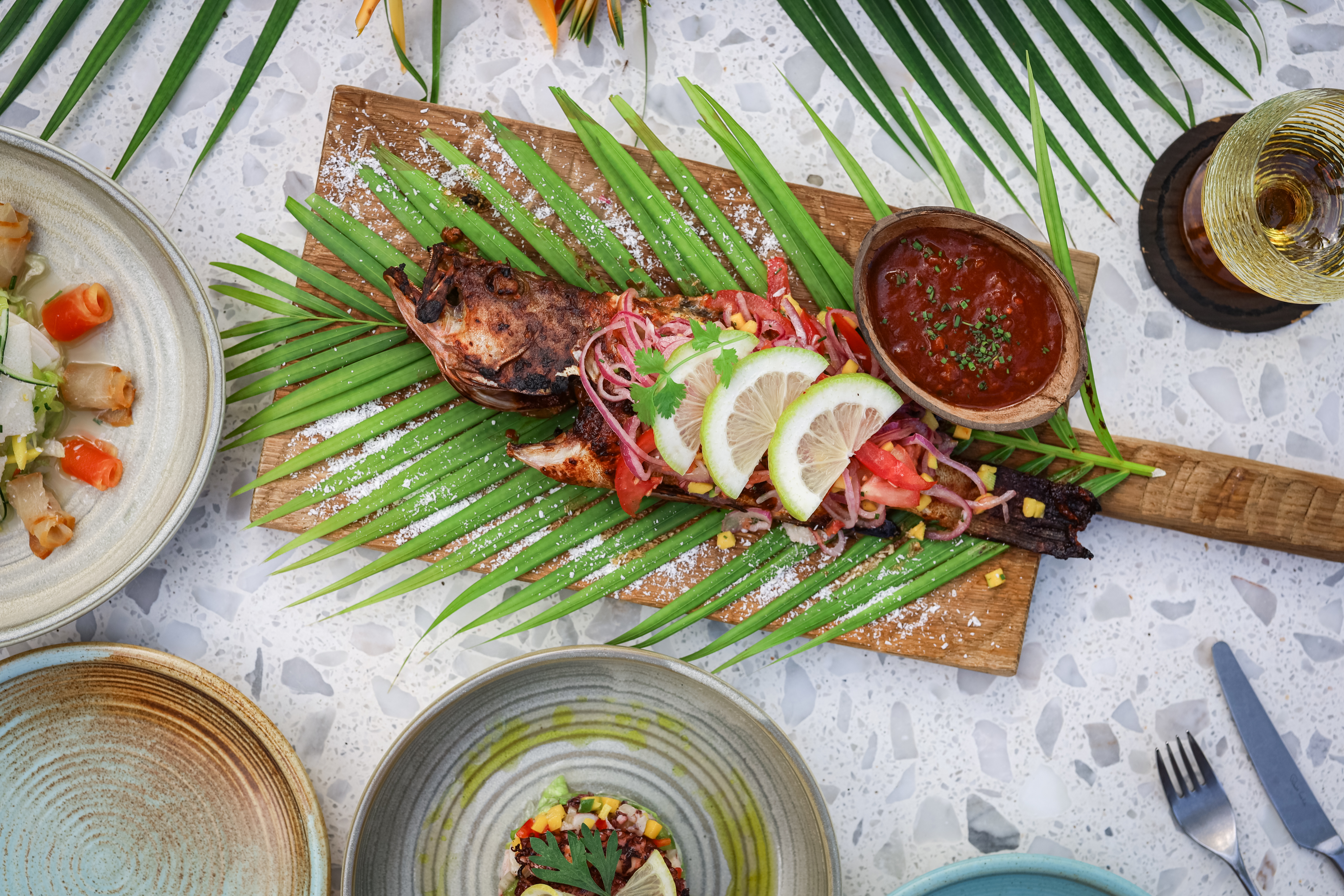 Whole grilled fish on a plank with fixings, a bowl of sauce, and lemon slices.